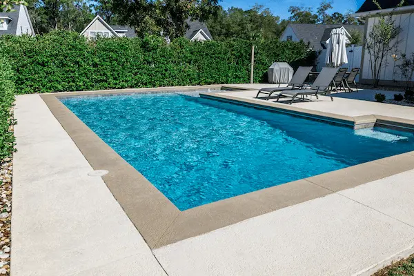 A rectangular new swimming pool with tan concrete edges in the fenced backyard of a new construction house with privacy hedges.